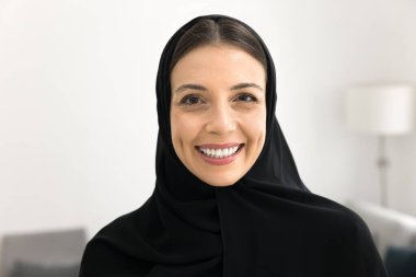 Siyah tesettürlü neşeli genç bir Arap kadın ve kadın Müslüman kumaşı kameraya bakıyor, gülümsüyor, görüntülü konferans için poz veriyor kafa fotoğrafı çekiyor, evde duruyor, gülüyor, ön planda.