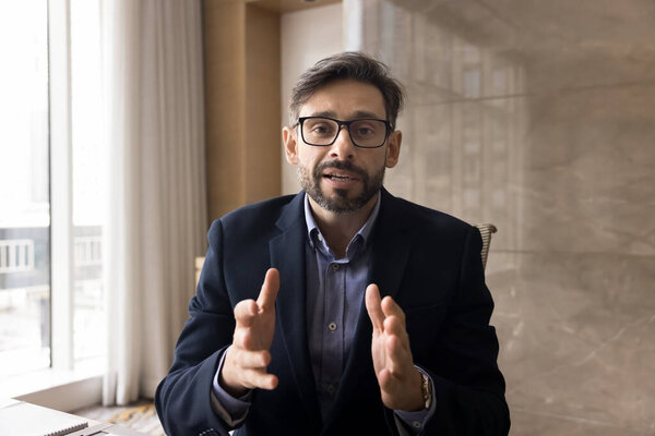 Serious Hispanic business professional man talking on video call from office, speaking on conference chat, looking at camera, moving hands, using business Internet communication. Head shot portrait