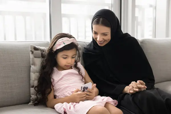 stock image Happy Muslim mom watching little daughter kid using mobile phone, playing game for children on Internet, using learning school application, typing on mobile phone, texting message