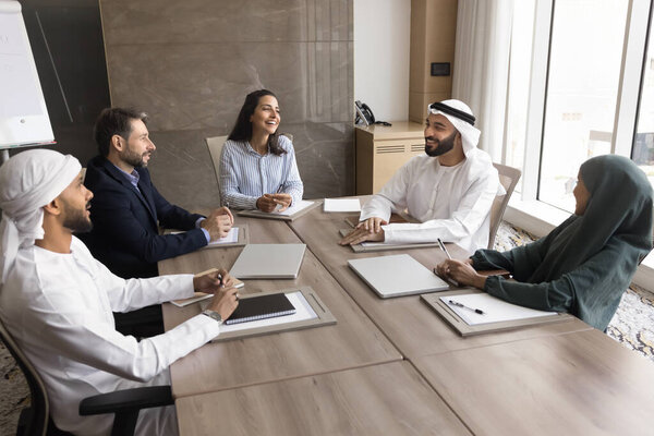 Positive diverse business team of Arabian and European entrepreneurs meeting for brainstorming, planning company management, talking, laughing. Multiethnic board of directors high angle shot