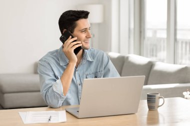 Akıllı telefondan konuşan mutlu, genç, İspanyol, iş yerinde dizüstü bilgisayarla oturuyor, pencereye bakıyor, gülümsüyor, telefondan keyif alıyor, sohbet ediyor.