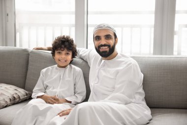 Mutlu, yakışıklı bir Arap baba ve beyaz Müslüman elbiseli sevimli bir oğlan birlikte kanepede oturuyor, kameraya bakıyor, gülümsüyor, gülüyor, aile fotoğrafı için poz veriyor.