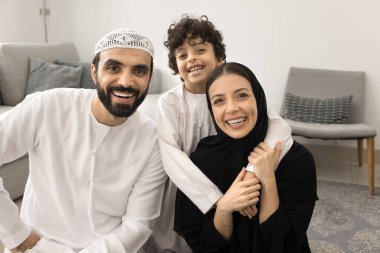 Mutlu Müslüman çocuk ve Arap elbiseli neşeli ebeveynler ev zemininde portre için poz veriyor, sarılıyor, gülüyor, dişlek tebessümle kameraya bakıyor. Anne tesettürlü çocuk, eğleniyor.
