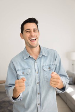 Mutlu genç Latin adam evde eğleniyor, apartman dairesinde dans ediyor, kazanan eller yapıyor, kazanan jest yapıyor, kameraya bakıyor, gülümsüyor, gülüyor, şarkı söylüyor. Gündelik dikey portre