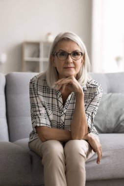 Gerçekten çekici, orta yaşlı bir kadın kameraya poz veriyor, evde dinleniyor, kanepede tek başına oturuyor, dikey portre çiziyor. Göz giyim mağazası, göz kontrolü hizmetleri reklamı.