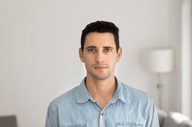 Ciddi yakışıklı genç Latin bir adam, arka planda beyaz bir duvarla evde ön kafaya poz veriyor, kameraya bakıyor. Çekici erkek, sıradan kot giyen, portre için içeride duran İspanyol manken.