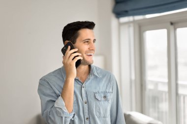 Neşeli, yakışıklı, İspanyol asıllı genç bir adam evde cep telefonuyla konuşuyor, telefon görüşmesinin tadını çıkarıyor, başka yere bakıyor, cep telefonuyla konuşuyor, gülümsüyor, gülüyor.