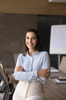 Kendine güvenen, zarif gömlekli genç İspanyol iş kadını büyük toplantı masasında portre için poz veriyor, kollarını kavuşturup kameraya bakıyor, güven jesti yapıyor, gülümsüyor. Dikey gömlek