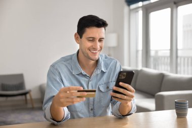 Finansal internet teknolojisinden zevk alan neşeli genç İspanyol müşteri, e-ticaret web hizmetleri için kredi kartıyla ödeme yapıyor, cep telefonu kullanıyor, gülümsüyor.