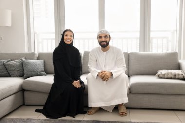 Müslüman kıyafetleri içinde mutlu bir Orta Doğulu çift evde kanepede oturuyor, kameraya bakıyor ve gülümsüyor. Olumlu Arap genç karı ve koca tam vücut aile portresi