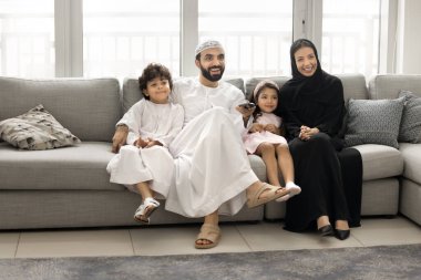 Mutlu Müslüman ebeveynler ve evde televizyon seyreden iki küçük kardeş kardeş birlikte rahat bir kanepede dinleniyorlar, sarılıyorlar, gülüyorlar, uzaktan kumandanın tuşlarına basıyorlar.