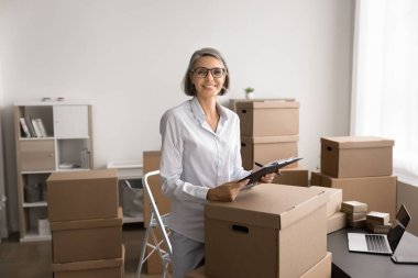 Olgun bir kadın, mukavva kutularla çevrili ofiste duruyor ve müşteriye gönderilmek üzere paketler hazırlıyor. Envanter yönetimi, sipariş tamamlama. Çevrimiçi satış, küçük işletme sahibi portresi