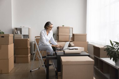 Olgun bir kadın, karton kutularla dolu bir odada iskemlede oturur, dizüstü bilgisayarla çalışır, envanter işlemleri, işlemler, paketleme ürünleri yönetiminde yer alır ve malları nakliyeye hazırlar.