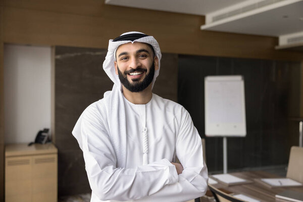 Happy confident Arabic company owner, executive man in white traditional Arabic clothes posing with hands crossed with white board behind, looking at camera with toothy smile for head shot portrait