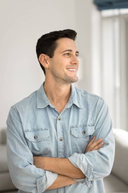 Düşünceli, çekici, rahat gömlekli, evde poz veren, elleri çaprazlanmış, güzel düşüncelere gülümseyen, hayal kuran, düşünceli genç Latin bir adam. Dikey portre