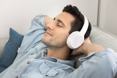 Kablosuz kulaklıklı mutlu genç Latin adam evde müzik dinliyor, rahatlamanın, ses eğlencesinin, çevrim içi oyuncunun çevresindeki müzik listesinin keyfini çıkarıyor, yakın gözlerle kanepeye yaslanıyor.