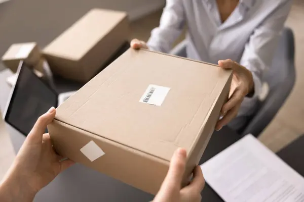 stock image Top above close up view unknown woman hands, postal office worker extending packaged cardboard parcel box towards marketplace client, female customer. Selling and buying, dispatch and shipment service