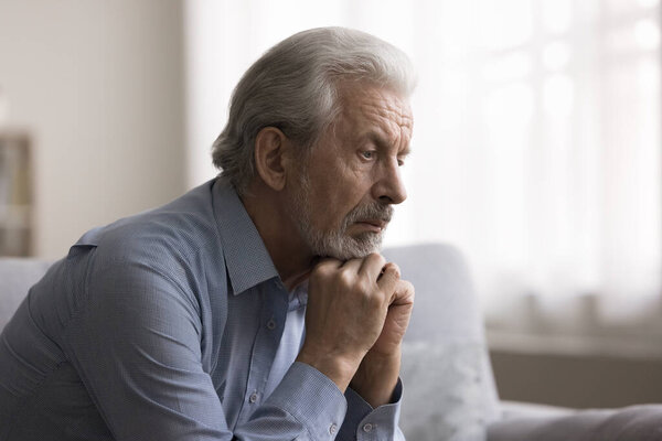Pensive older grey-haired man put chin on hands, sit alone on sofa engrossed in deep sad thoughts, suffering from loneliness, feel abandoned, having apathetic or nostalgic mood, coping with problems