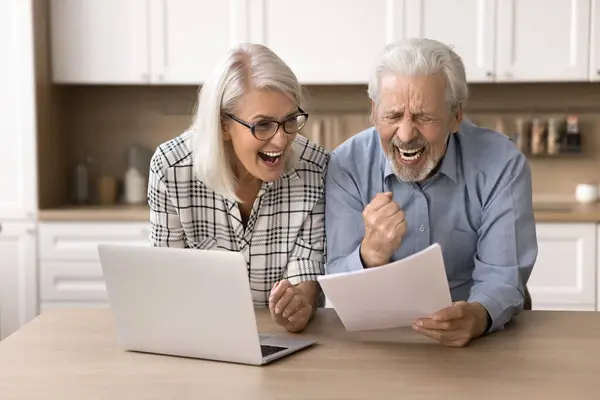 stock image Happy aged couple read letter, hold document, bank statement, feels excited by great news, receive notice of loan repayment, money back, gesturing with joy. Celebrate financial success, pension growth