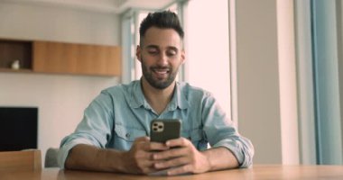 Mutlu Latin serbest iş adamı akıllı telefonu ev iş masasında kullanıyor, cep telefonuyla yazı yazıyor, internet teknolojisinden keyif alıyor, online iletişim kuruyor, sohbet ediyor, gülümsüyor, gülüyor.