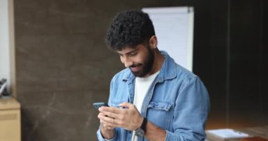 Neşeli genç Arap girişimci günlük giysiler içinde İnternet teknolojisini iş iletişimi için kullanıyor, ofisteki cep telefonuna mesaj yazıyor, dijital cihaz tutuyor, gülümsüyor.