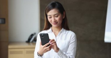 Mutlu Asyalı çalışan kız cep telefonunda mesaj yazıyor, internette geziniyor, gülümsüyor, internet teknolojisinden zevk alıyor, yüzünü çeviriyor, online iletişimi düşünüyor.