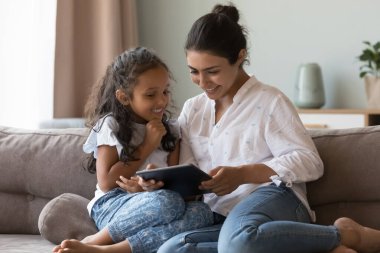 Genç Hintli anne, küçük kızına dijital tablette yeni bir uygulama gösteriyor. Aile, modern kablosuz cihazla evde vakit geçiriyor, internette eğleniyor. Z jenerasyonu teknoloji kavramını kullan