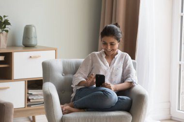 Genç Hintli kadın akıllı telefonuyla koltukta dinleniyor hafta sonunu internette yalnız geçiriyor, gülümsüyor sosyal medyada sohbet ediyor, ürünleri seçiyor, e-ticaret perakende uygulaması kullanarak hizmet satın alıyor.