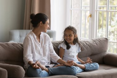 Genç Hintli kadının tatlı kızı parmaklarını kavuşturarak Mudra jestini kanepeye oturup meditasyon yaparken kahkahalar attırıyor. Ebeveynler çocuk yoga ve iyi yaşam alışkanlıkları, yaşam tarzı konsepti öğretir