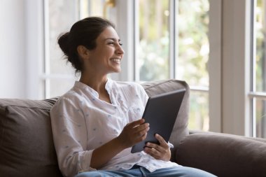 Mutlu Hintli kadın oturma odasındaki kanepede gülüyor kablosuz dijital tablet cihazıyla internette eğleniyor. E-ticaret hizmetleri istemcisi, basit uzak düzen, uygulama ve İnternet kullanımı kavramı