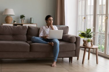 Konforlu oturma odasında düşünceli Hintli bir kadın kanepeye oturur, dizüstü bilgisayar kullanır, yan gözle bakar, internet alışverişini düşünür, e-alışveriş yapar, tele-iş yapar, çevrimiçi çalışırken çözüm üzerinde düşünür. Teknoloji kavramı