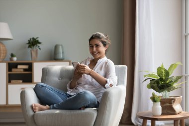Çekici genç Hintli kadın rahat koltukta rahatlıyor ve kamerada akıllı telefon gülümsemesi pozu veriyor. Modern teknoloji kullanıcısı, konfor hayatı için yeni mobil uygulama, elektronik alışveriş ve hizmet alıcısı portresi konsepti