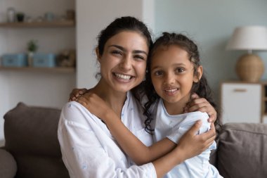 Gülümseyen küçük kız, genç annesine sarılıyor koltukta oturmuş kameraya poz veriyor. Güzel Hintli mutlu aile portresi, annelik ve velayet, koşulsuz aşk konsepti.