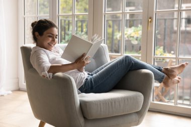 25 'lerinde gülümseyen Hintli bir kadın kitap okuyor rahat bir odada rahat koltukta dinleniyor, boş zamanlarını kitapçıdan alınan favori edebiyatın tadını çıkarıyor. Kitapseverlerin evde zaman geçirmesi, hobi konsepti