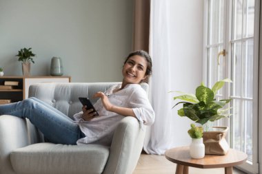 Sevimli Hintli kadın koltukta rahatla gülümse ve akıllı telefon kullan. Çevrimiçi mal alıcısı, sosyal medya ya da rahat yaşam için yeni mobil uygulama kullanımı. Elektronik ticaret hizmetleri kullanıcısı, modern teknoloji konsepti