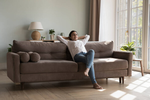 Inside clean cozy light living room young Indian woman resting on sofa put hands behind head closed eyes enjoy day off and fresh conditioned air at modern house. Relaxation, weekend, leisure concept