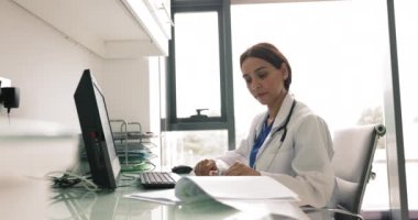 Genç 35 'lik kadın doktor masa başında oturur, hasta kayıtlarını inceler, tıbbi geçmişi, test sonuçlarını ve notları öğrenir, yazılı bilgileri analiz eder, tedavi planı üzerinde düşünür, reçete yazar.