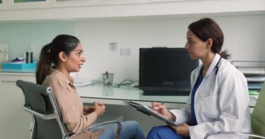 Terapistle konuşan genç Hintli kadın, özel klinikteki son semptomları açıkla. İlgili sorular soran kadın doktor, hasta sağlık şikayetleri, endişeler ve notlar alan.