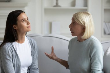 Strict anxious mature mother lecturing scolding annoyed grown daughter, sitting on sofa at home, parent and child quarrel, generations conflict, stubborn young woman ignoring angry senior mum clipart