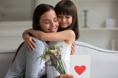 Gülümseyen anne, sevimli küçük kızını kapalı gözlerle kucaklıyor, hediye ve tebrik için teşekkür ediyor, kalpli ve çiçekli tebrik kartı, anne doğum gününü kutlayan aile, 8 Mart ya da anneler günü