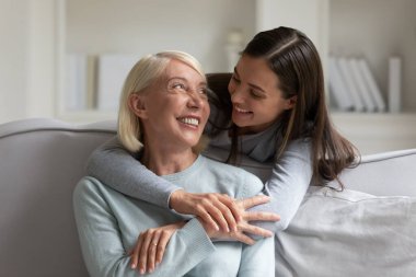 Gülümseyen torun, olgun büyükanneye arkadan sarılıyor, evde koltukta oturuyor, aile birlikte güzel anlar yaşıyor, yetişkin kız ve büyük anne birbirine bakıyor, iki nesil boyunca.