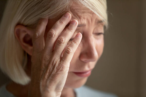 Close up unhappy mature woman crying, thinking about problems, frustrated older female suffering from strong headache or migraine, touching forehead, feeling unwell, dementia or mental disorder