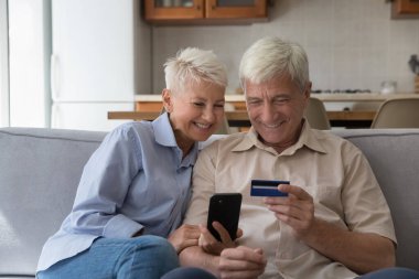 Mutlu yaşlı çift evdeki koltukta oturup akıllı telefon ve kredi kartıyla online alışverişin tadını çıkarıyor. Olgun eşler elektronik alışveriş yapar, güvenli ödeme yapar, ani para transferi yapar, ekommerce müşterileri