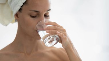 Duştan sonra havluyla gelen genç kadın susuzluğunu hisset ev banyosunda bardaktan temiz maden suyu iç, kız temiz kristal akının tadını çıkar, sağlıklı yaşam tarzını takip et, diyet yap, refah, hidrasyon kavramı.