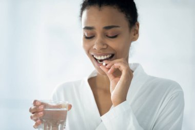Gülümseyen melez genç bir kadın günlük vitamin veya takviyelerini banyoda bir bardak suyla alıyor, mutlu Afrikalı bir Amerikalı kız ilaç ya da uyuşturucu alıyor, evde banyo yaparken doğum kontrol hapı içiyor.