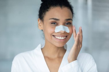 Gülümseyen Afrikalı Amerikalı genç kadının portresi burun bantlarını temizleme gözenekleri ya da siyah noktalar, mutlu melez kadınlar banyo ve cilt konsepti için yüz işlemleri yapıyor.