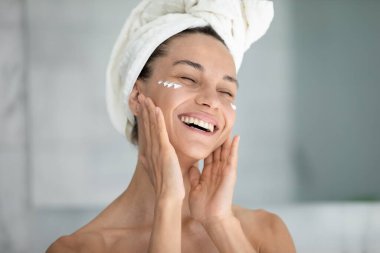 Duştan sonra banyodaki neşeli genç kadın nemlendirici yüz kremi ya da maske sür. Gülümseyen kadın, sabahları banyo ve cilt bakımında günlük güzellik prosedürleri uygula.