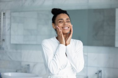 Bornoz giymiş mutlu Afro-Amerikalı genç bir kadın dokunmatik masaj, ameliyattan sonra parlayan pürüzsüz bir yüz, gülümseyen melez bir kadın güzellik tedavisinden memnun, cilt konsepti