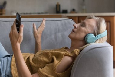 Kaygısız, barışçıl yaşlı bir kadın kanepeye uzanmış akıllı telefonuyla müzik listesini dinliyor, kaliteli sesin tadını çıkarıyor, dijital yayın yapan çevrimiçi hizmetler sunuyor. Evde vakit geçirmek, yaşlı insan konsepti hobisi.