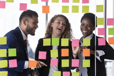 Çok mutlu, çok ırklı üç iş arkadaşı modern ofiste birlikte çalışıyor. Yapışkan notları bilgiyle hazırlıyor, fikirler kuruyor, iş stratejileri kuruyor, cam duvara iliştirilmiş görevler yapıyor. Takım çalışması konsepti